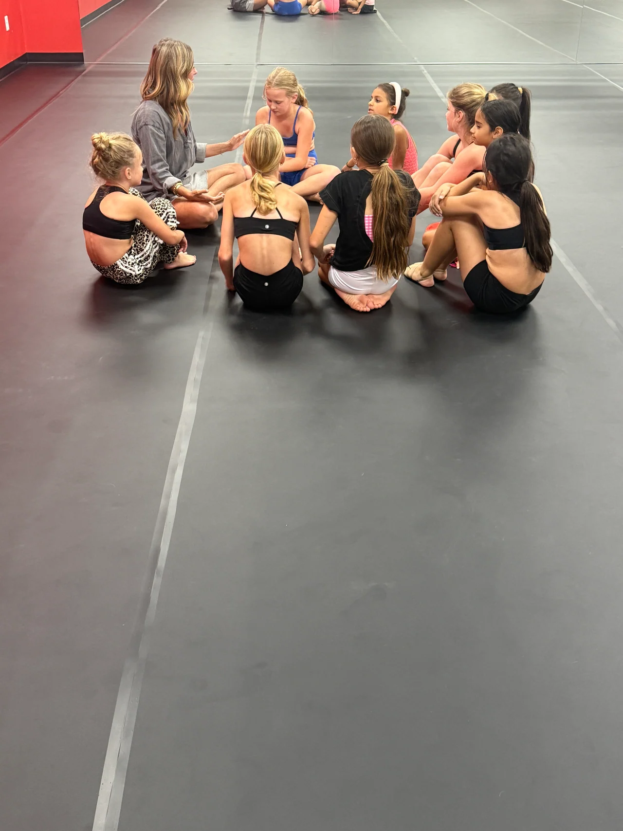 A group of young female dancers in dance attire sitting in a semi-circle on the dark studio floor, listening to their instructor.